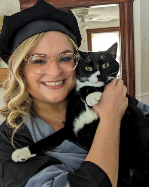 A smiling woman with blond hair, wearing glasses and her cap and gown, holding her black and white cat